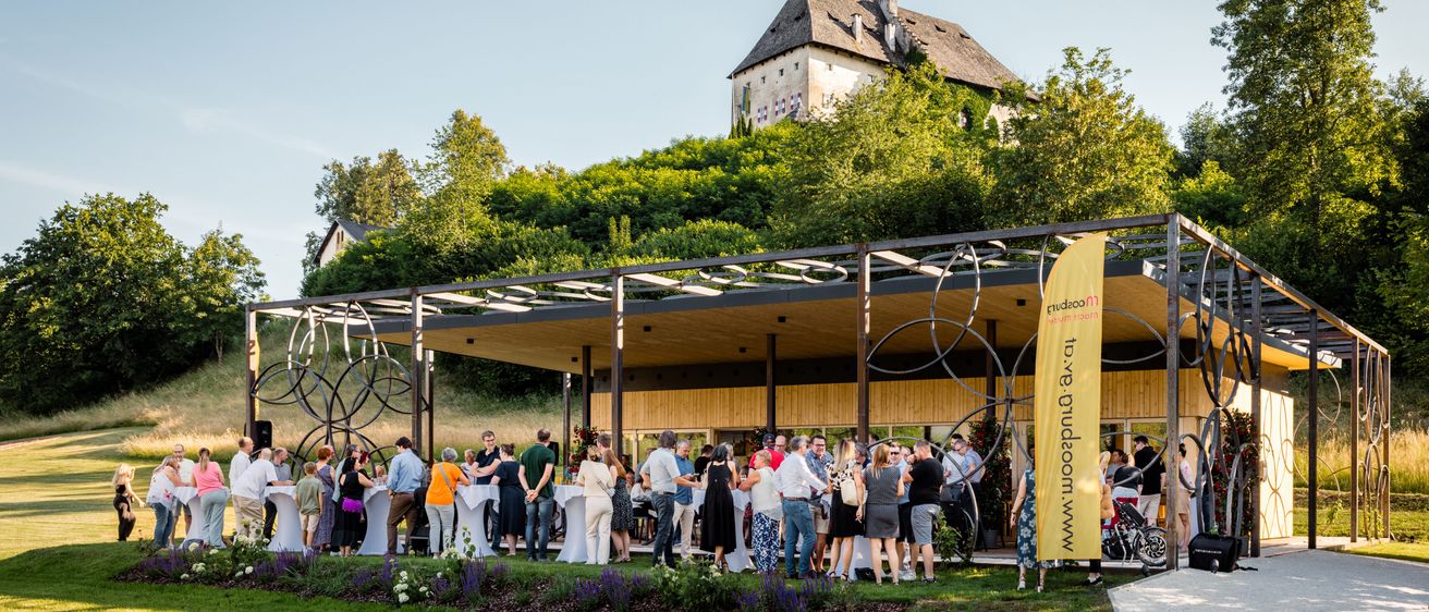 Menschen versammeln sich in einem Pavillon unter einem Schloss auf einem Hügel, umgeben von Grün, unter einem klaren Himmel.