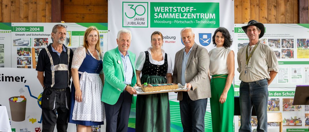 Eine Gruppe von Menschen steht lächelnd auf einer Bühne für die Kamera. Eine Frau in traditioneller Kleidung hält einen Kuchen. Hinter ihnen befindet sich ein Banner mit Text und Logos.