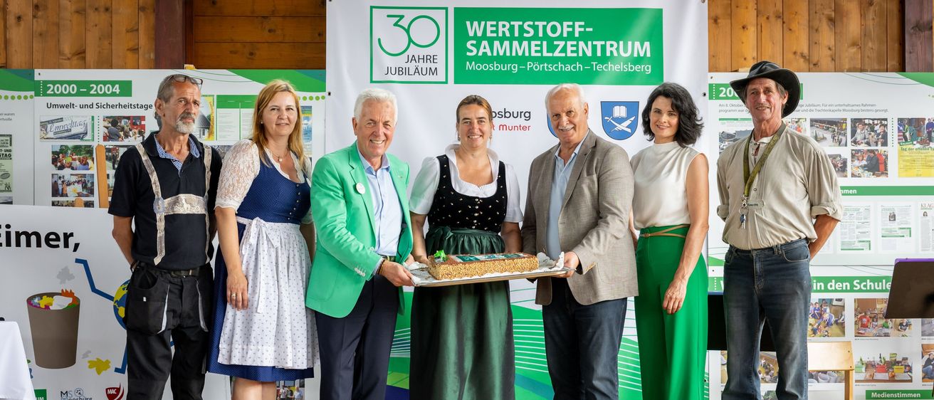 Eine Gruppe von Menschen steht lächelnd auf einer Bühne für die Kamera. Eine Frau in traditioneller Kleidung hält einen Kuchen. Hinter ihnen befindet sich ein Banner mit Text und Logos.