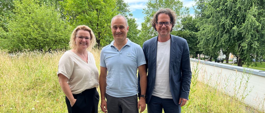Drei Menschen stehen in einem Feld mit hohem Gras. Eine Frau links und zwei Männer. Sie lächeln und posieren für ein Foto. Hinter ihnen sind Bäume und eine Mauer.