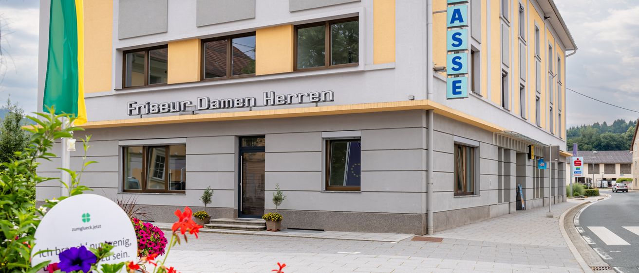Ein Gebäude mit einem Schild, das Friseur Damen Herren besagt. Es hat einen gefliesten Eingang, Topfpflanzen und Blumen davor.