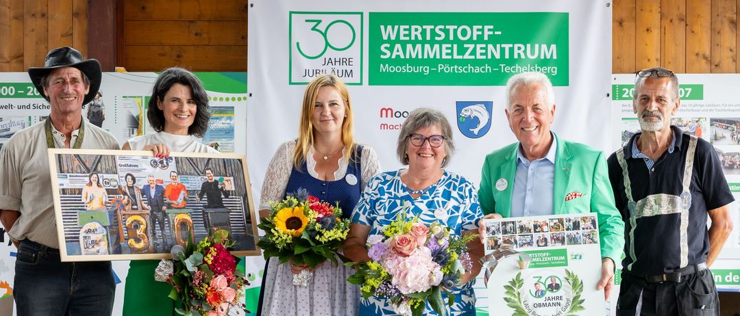 Eine Gruppe von Menschen steht auf einer Bühne mit Blumen und einem Banner, das 'Herzlich willkommen 30 Jahre Wertstoff-Sammelzentrum Moosburg-Portschach-Tebelsberg' besagt. Sie lächeln und halten ein Plakat und Zertifikat.