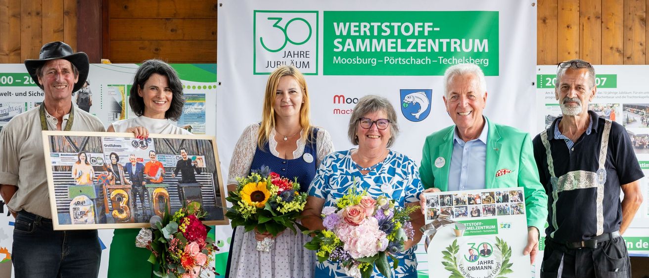 Eine Gruppe von Menschen steht auf einer Bühne mit Blumen und einem Banner, das 'Herzlich willkommen 30 Jahre Wertstoff-Sammelzentrum Moosburg-Portschach-Tebelsberg' besagt. Sie lächeln und halten ein Plakat und Zertifikat.
