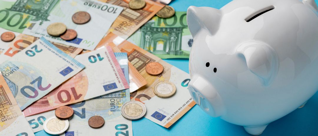 Eine weiße Sparschweinbank mit Münzeinwurf steht auf einem blauen Hintergrund, umgeben von verschiedenen Euro-Banknoten und Münzen.