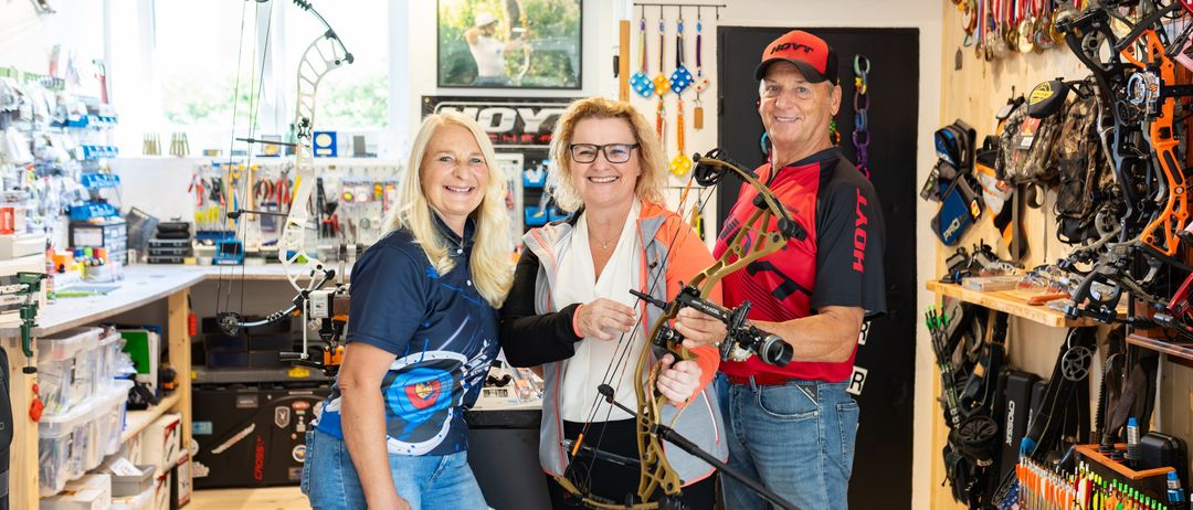 Drei Personen posieren mit einem Bogen in einem Geschäft. Ein Mann in Rot und Schwarz hält den Bogen, während zwei Frauen, eine in Blau und eine in Weiß, neben ihm stehen. Das Geschäft hat verschiedene Ausrüstungsgegenstände und ein Schild mit der Aufschrift 'HOYT ARCHERY'.