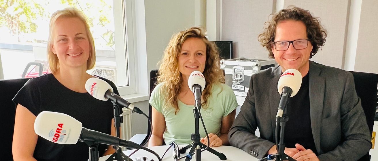 Drei Personen sitzen in einem Studio um einen Tisch, jeder mit einem Mikrofon davor. Zwei Frauen, eine in Schwarz, die andere in Grün, und ein Mann im Anzug. Papiere und Kabel auf dem Tisch. Fenster und Bäume im Hintergrund sichtbar.