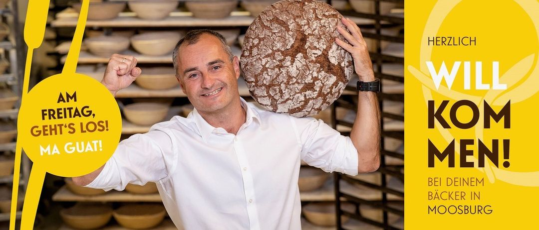 Ein Mann in einem weißen Hemd hält ein großes, rustikal aussehendes Brot vor seiner Brust und lächelt in die Kamera.
