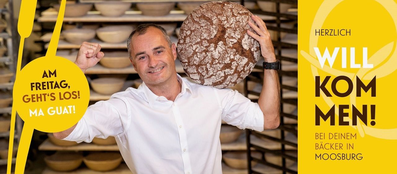 Ein Mann in einem weißen Hemd hält ein großes, rustikal aussehendes Brot vor seiner Brust und lächelt in die Kamera.