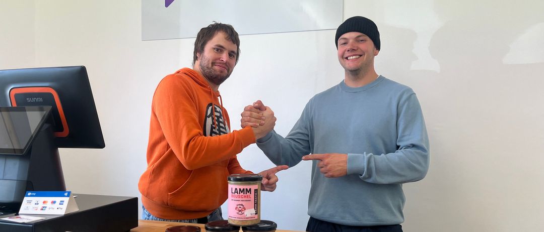 Zwei Männer stehen hinter einem Tresen, einer zeigt auf einen Topf LamM Ragout und der andere schüttelt Hände. Hinter ihnen befindet sich ein Banner mit dem Text 'Rainer'.