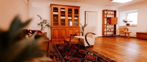 Ein Wohnzimmer mit Holzboden, einem Holzschrank, einem Teppich, einem Sofa, einem Stuhl und einer Topfpflanze. Es gibt auch ein Regal mit einer Lampe.