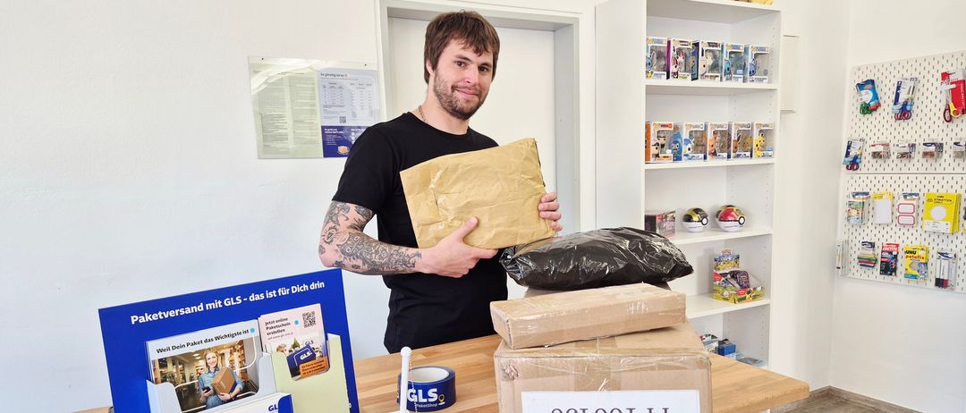 Ein Mann mit Tattoos steht hinter einem Schreibtisch und hält ein Paket. Der Schreibtisch hat Kisten, Klebeband und GLS-Informationen. Dahinter steht ein Regal mit Pokemon-Figuren und eine Pinnwand.