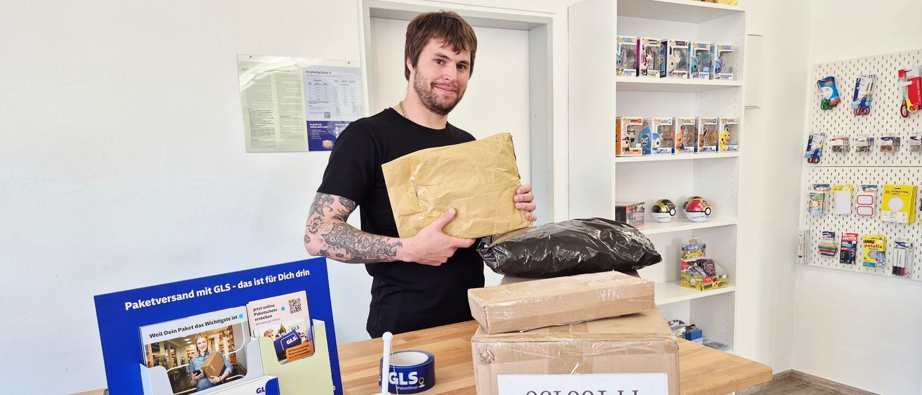 Ein Mann mit Tattoos steht hinter einem Schreibtisch und hält ein Paket. Der Schreibtisch hat Kisten, Klebeband und GLS-Informationen. Dahinter steht ein Regal mit Pokemon-Figuren und eine Pinnwand.