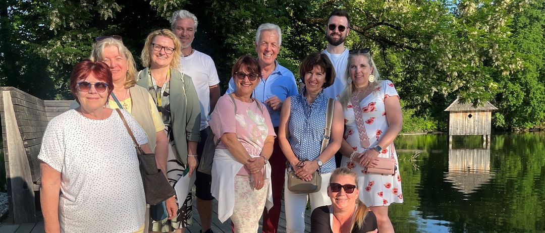 Eine Gruppe von Erwachsenen, einschließlich Männer und Frauen, posiert für ein Foto am See. Sie tragen Freizeitkleidung und einige tragen Sonnenbrillen. Sie stehen auf einem Holzsteg.