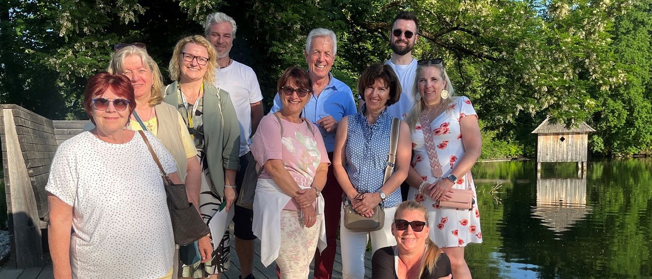 Eine Gruppe von Erwachsenen, einschließlich Männer und Frauen, posiert für ein Foto am See. Sie tragen Freizeitkleidung und einige tragen Sonnenbrillen. Sie stehen auf einem Holzsteg.