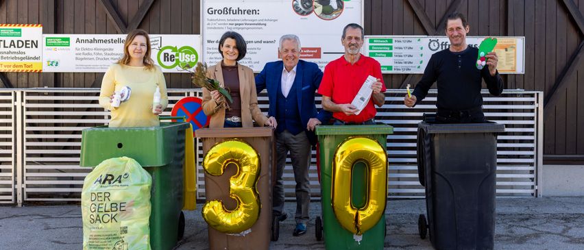 Fünf Personen stehen vor Recycling-Tonnen, jede mit der Aufschrift 30. Sie lächeln und posieren für ein Foto. Dahinter ist ein Schild mit den Texten 'Wir ubernehmen' und 'Re-Use' zu sehen.