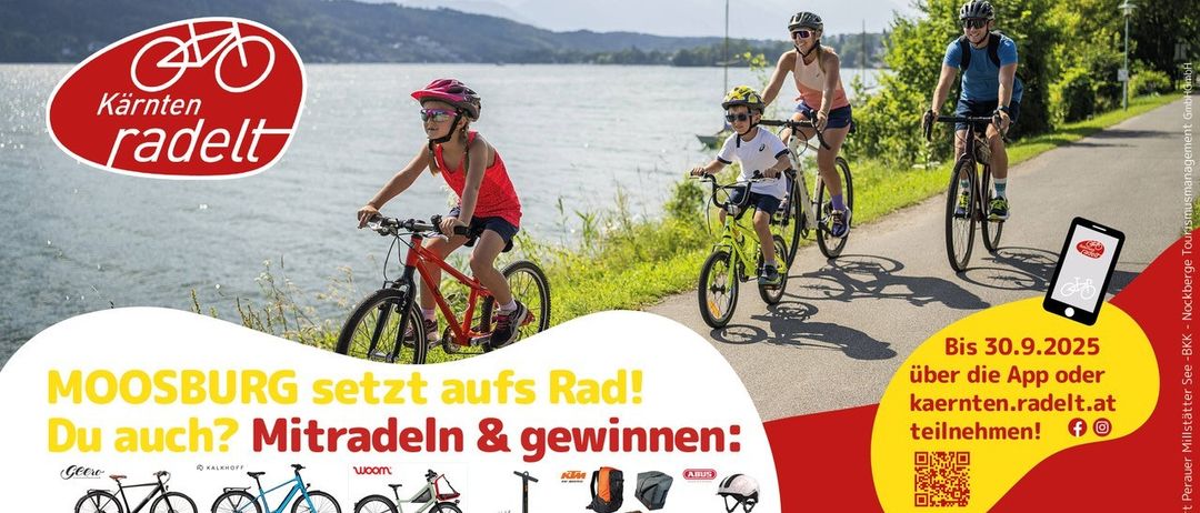 Vier Radfahrer, darunter zwei Kinder, fahren auf einem Pfad am Seeufer. Sie tragen Helme und Sonnenbrillen. Im Hintergrund sind ein See und Berge zu sehen.