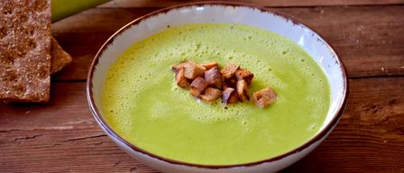 Eine Schüssel grüner Suppe mit Croutons auf einer Holzoberfläche, ein grünes Gemüse daneben.