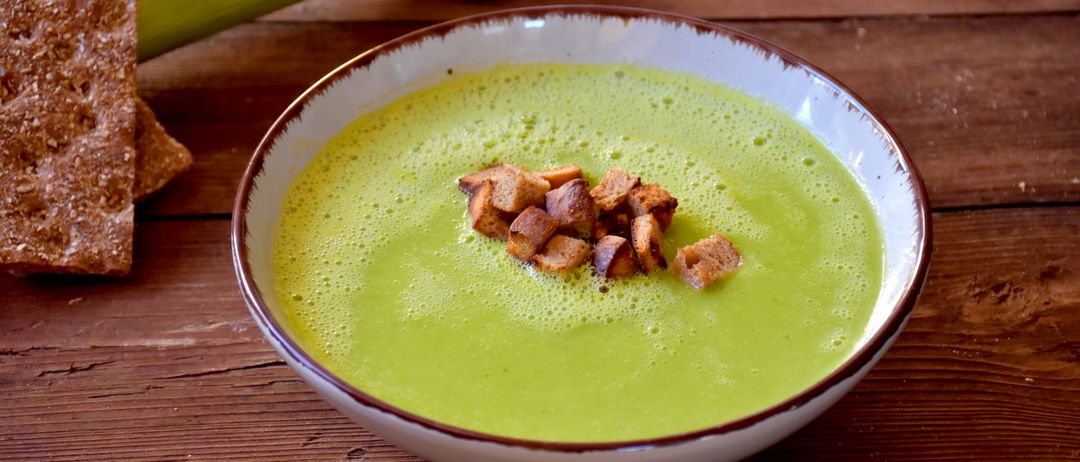 Eine Schüssel grüner Suppe mit Croutons auf einer Holzoberfläche, ein grünes Gemüse daneben.