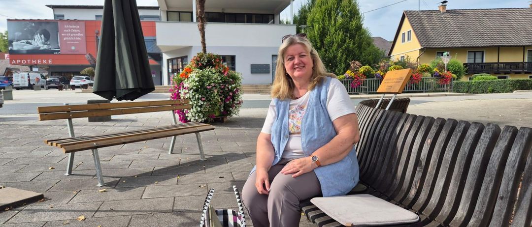 Eine Frau sitzt auf einer Bank in einem Stadtplatz. Ein Beutel liegt vor ihr. Hinter ihr befinden sich Gebäude, Bäume und ein geschlossener Schirm.
