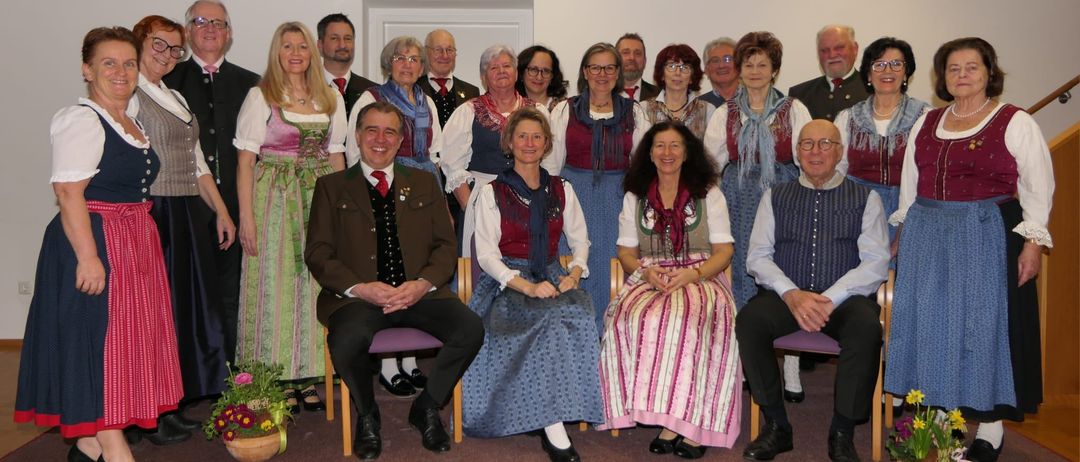 Eine Gruppe von Menschen, einige in traditioneller Kleidung, posiert für ein Foto in einem Raum mit weißer Wand und Holzboden, lächelnd.