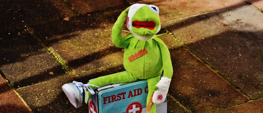 Ein Kermit der Frosch Plüsch sitzt auf einer Erste-Hilfe-Kiste. Er hat Verbände und einen roten Fleck im Gesicht.