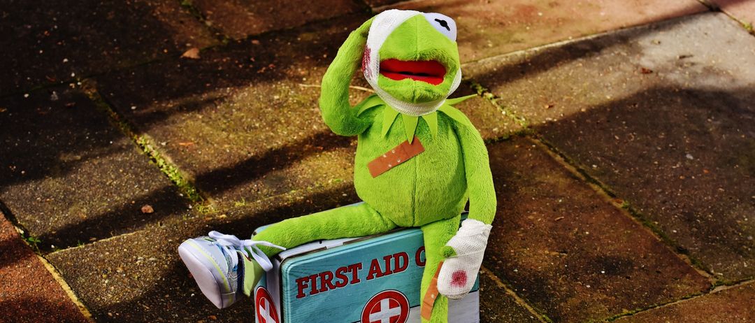 Ein Kermit der Frosch Plüsch sitzt auf einer Erste-Hilfe-Kiste. Er hat Verbände und einen roten Fleck im Gesicht.