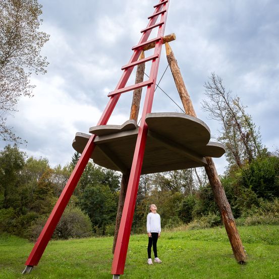 Ein junges Mädchen steht vor einer großen roten Leiter-artigen Struktur mit einer Betonplattform oben, umgeben von einem Grasfeld und Bäumen.