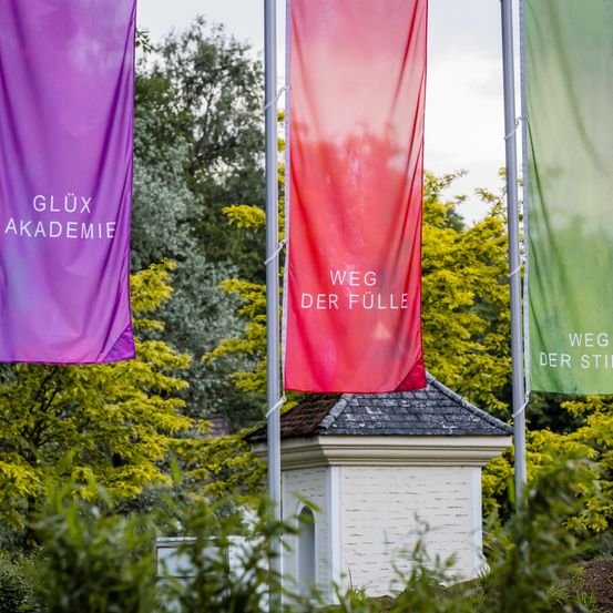 Drei Banner mit Wörtern in verschiedenen Sprachen stehen vor einem kleinen Gebäude auf Stangen. Der linke Banner ist lila mit weißen Text. Der mittlere Banner ist rot mit weißem Text. Der rechte Banner ist grün mit weißem Text.