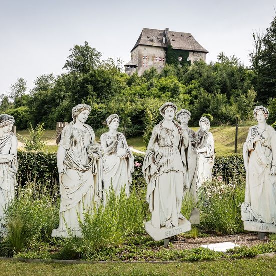 Sieben Statuen stehen in einem Garten mit grünen Büschen und Blumen, ein Schloss auf einem Hügel im Hintergrund.