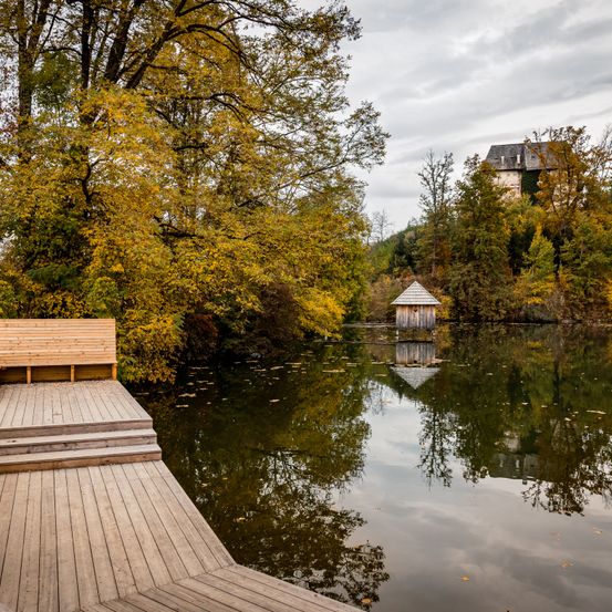 Eine Holzterrasse mit Stufen, die zu einem ruhigen See führen. Das Wasser ist ruhig und spiegelt Bäume und eine kleine Hütte wider. In der Ferne steht ein größeres Gebäude auf einem Hügel.