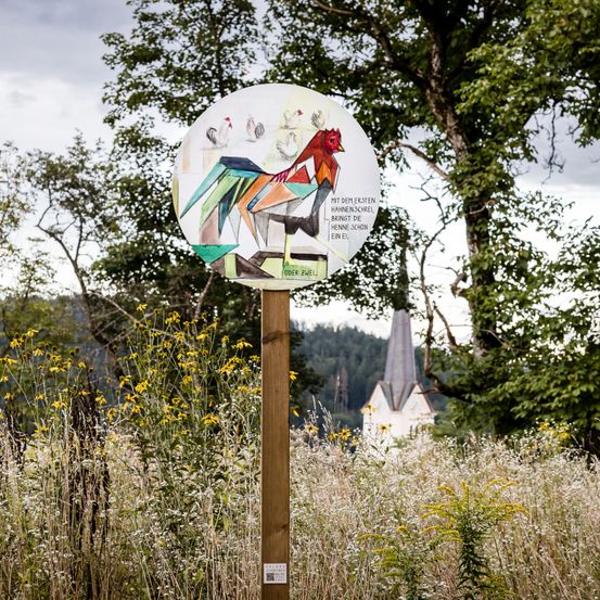 Ein farbenfrohes Bild eines Hahns auf einem Holzpfosten steht in einem Grasfeld mit Bäumen und einer Kirche im Hintergrund.