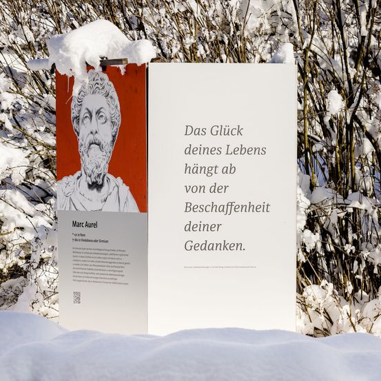 Ein Poster mit einem Bild von Marc Aurel steht draußen im Schnee. Es enthält einen Zitat von ihm, das lautet 'Das Glück deines Lebens hängt von der Beschaffenheit deiner Gedanken ab.'