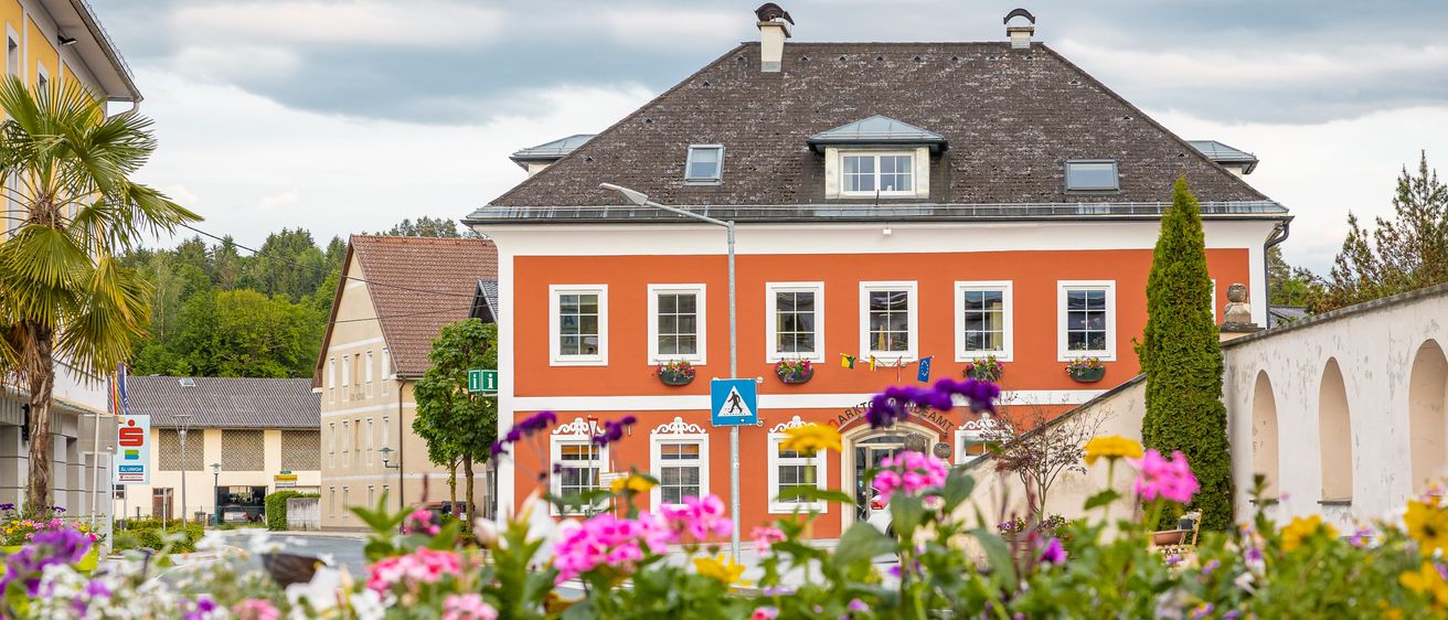Ein leuchtend orangefarbenes Gebäude mit weißen Fensterrahmen und mehreren Fenstern sowie Blumen in den Fensterkästen wird von einem hölzernen Pflanzgefäß mit bunten Blumen aus gesehen.