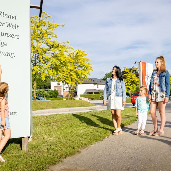Drei Frauen und zwei junge Mädchen gehen auf einem Pfad mit einem grünen Grasbereich. Ein Schild sagt 'Kinder sind unsere Zukunft'. Dahinter steht ein Baum mit gelben Blättern, Häusern und einem geparkten Auto.