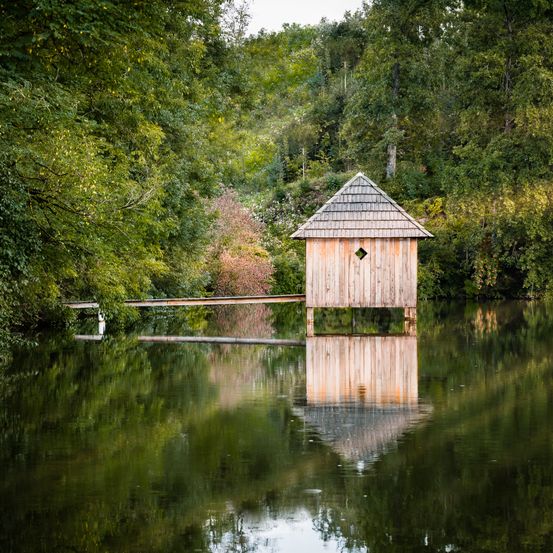 Eine kleine Holzhütte steht auf Stelzen in einem ruhigen See, umgeben von üppigem Grün. Die Spiegelungen der Hütte und der Bäume tanzen auf der Wasseroberfläche.