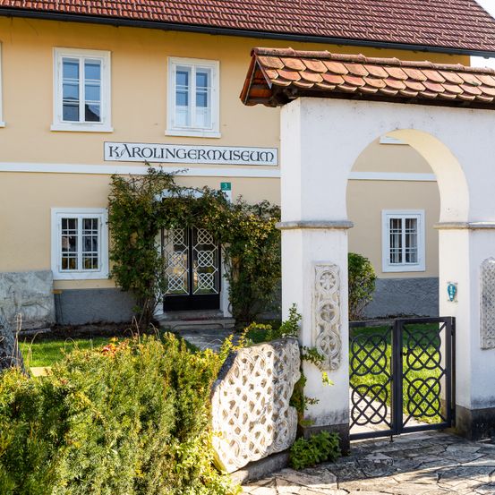 Ein gelbes Gebäude mit weißen Fenstern und mehreren Fenstern, benannt Karolingermuseum, hat ein braunes Ziegeldach und einen gewölbten Eingang mit einem dekorativen Tor.