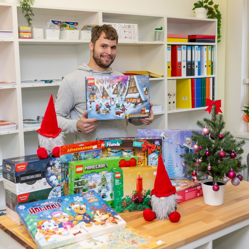 Ein Mann, der ein Lego-Set hält und von Spielzeug, Kisten, einem Weihnachtsbaum und einem Bücherregal umgeben ist.