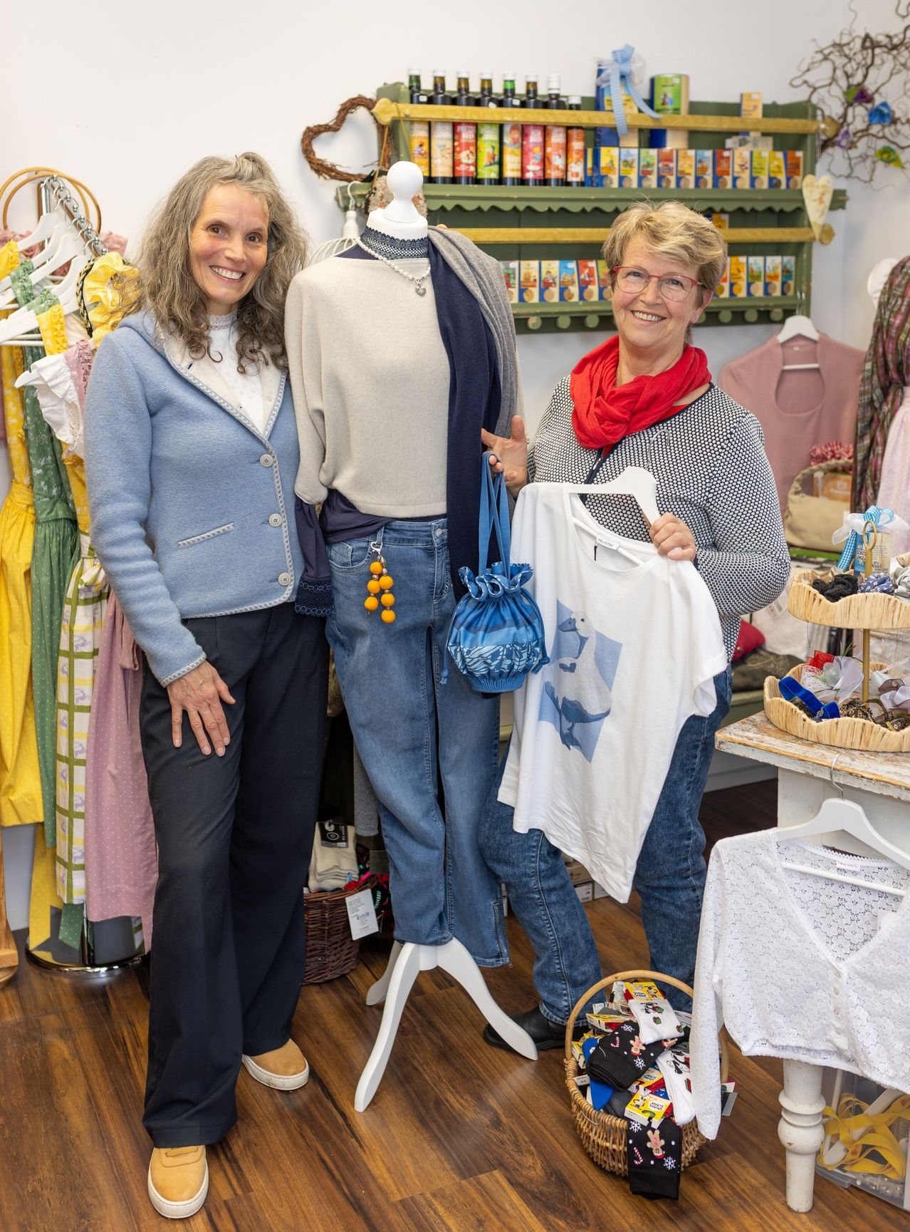 Zwei Frauen in einem Bekleidungsladen posieren für ein Foto. Eine hält ein weißes T-Shirt, während die andere neben einem Mannequin steht. Verschiedene Kleidungsstücke hängen im Hintergrund.