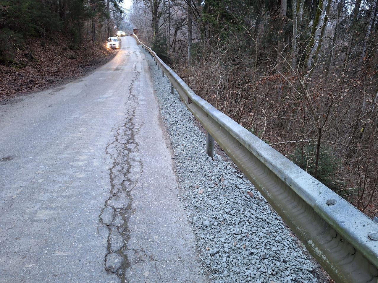 Eine Straße mit Schotter daneben und vorbeifahrenden Autos. Es gibt eine Leitplanke und Bäume ohne Blätter.
