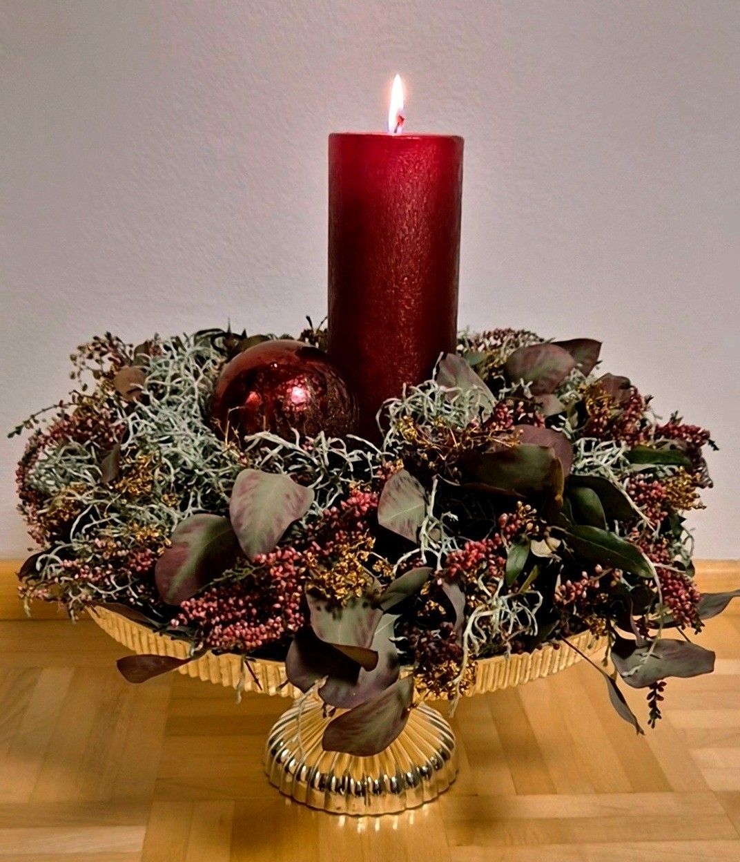 Eine rote Kerze steht auf einem goldenen dekorativen Zentrum, umgeben von Grünpflanzen und einer roten Kugel. Die Szene ist vor einem schlichten weißen Hintergrund angeordnet.