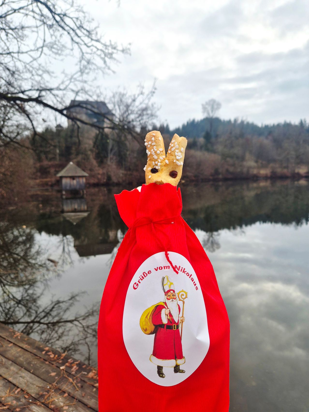 Ein roter Beutel mit einem Lebkuchenmann und einer Zeichnung von Santa Claus steht vor einem See. Im Hintergrund befindet sich ein Haus und eine Burg.