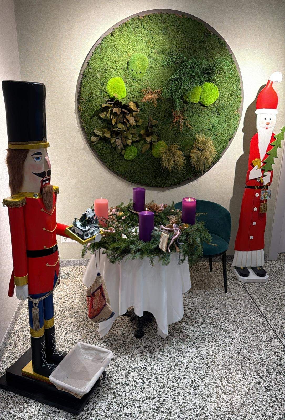 Ein Weihnachtsnussknacker hält eine Deko-Schale neben einem Tisch mit Kerzen, in der Nähe eines grünen Stuhls und einer großen Mooswand. Der Weihnachtsmann steht neben einem Spiegel.