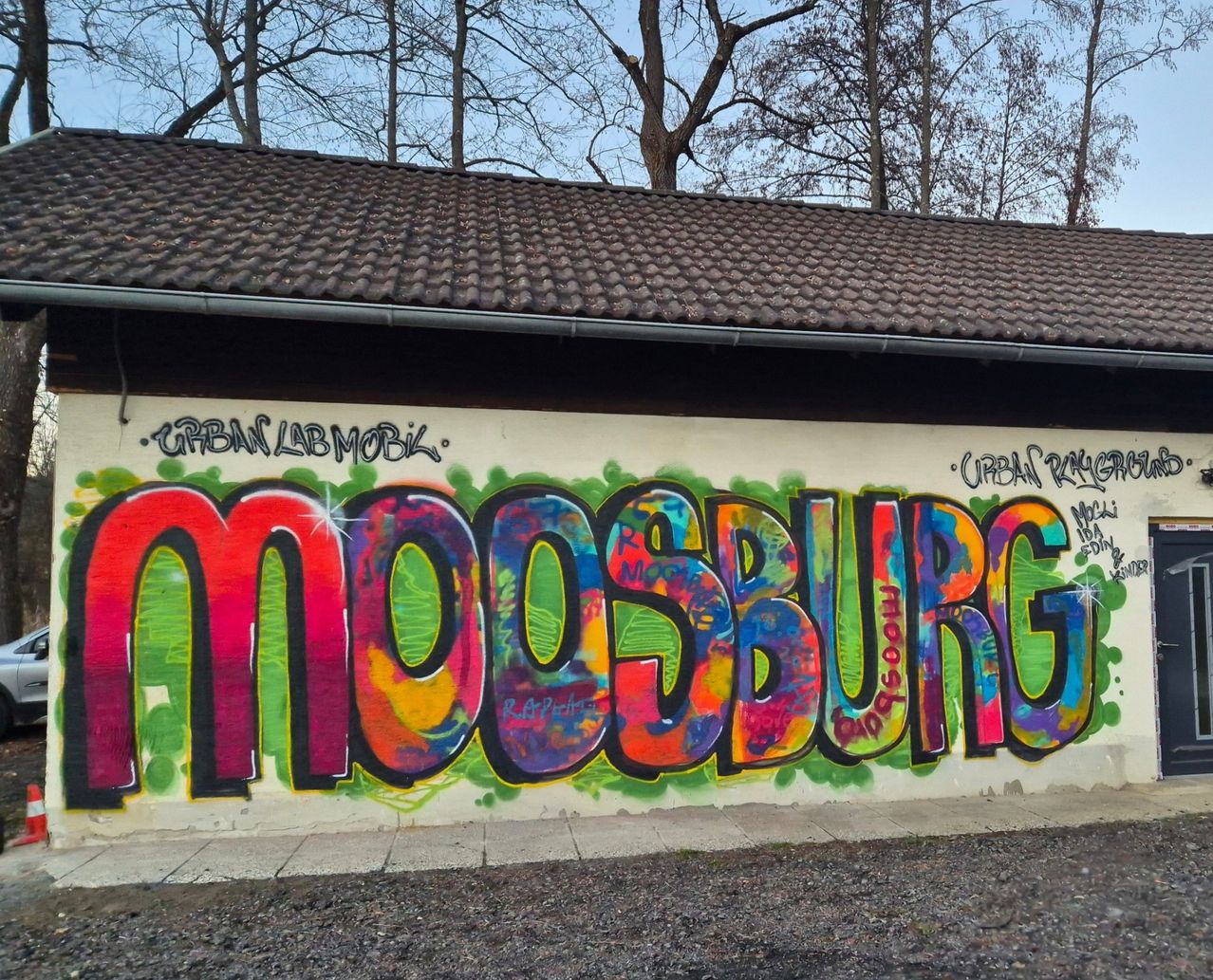 Graffiti an einer Gebäudewand mit dem Schriftzug 'Moosburg' in bunten Buchstaben. Bäume und ein Dach im Hintergrund.
