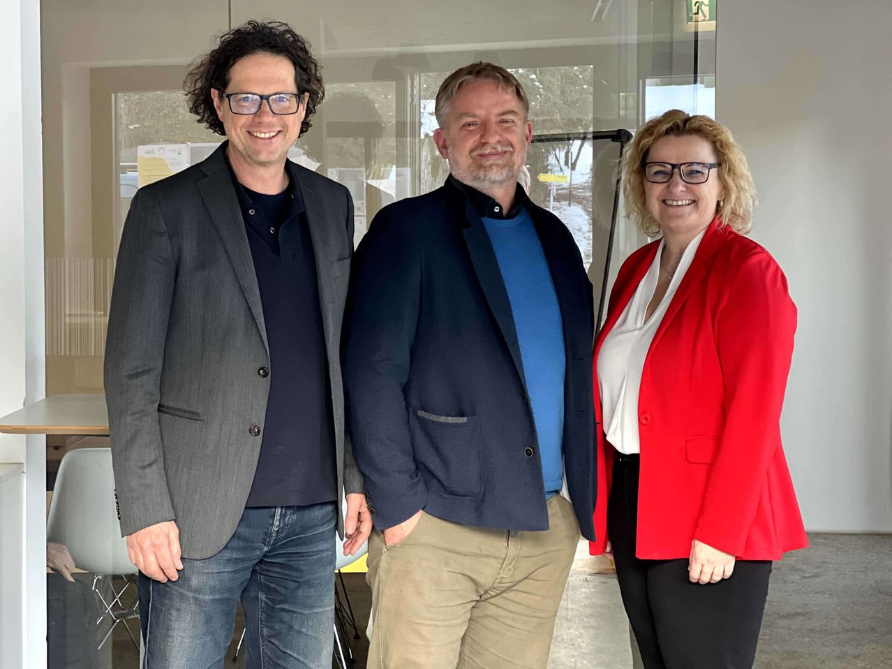 Drei Personen, die lächelnd vor einer Glaswand stehen, mit Reflexionen von Bäumen und einem Schild.