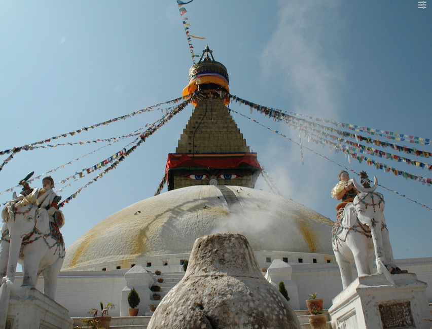 Der Bodhnath-Stupa, eine große weiße Kuppel mit Flaggen und einem rotäugigen Gesicht, hat zwei Elefanten mit Menschen davor. Davor stehen Pflanzen und Bäume.