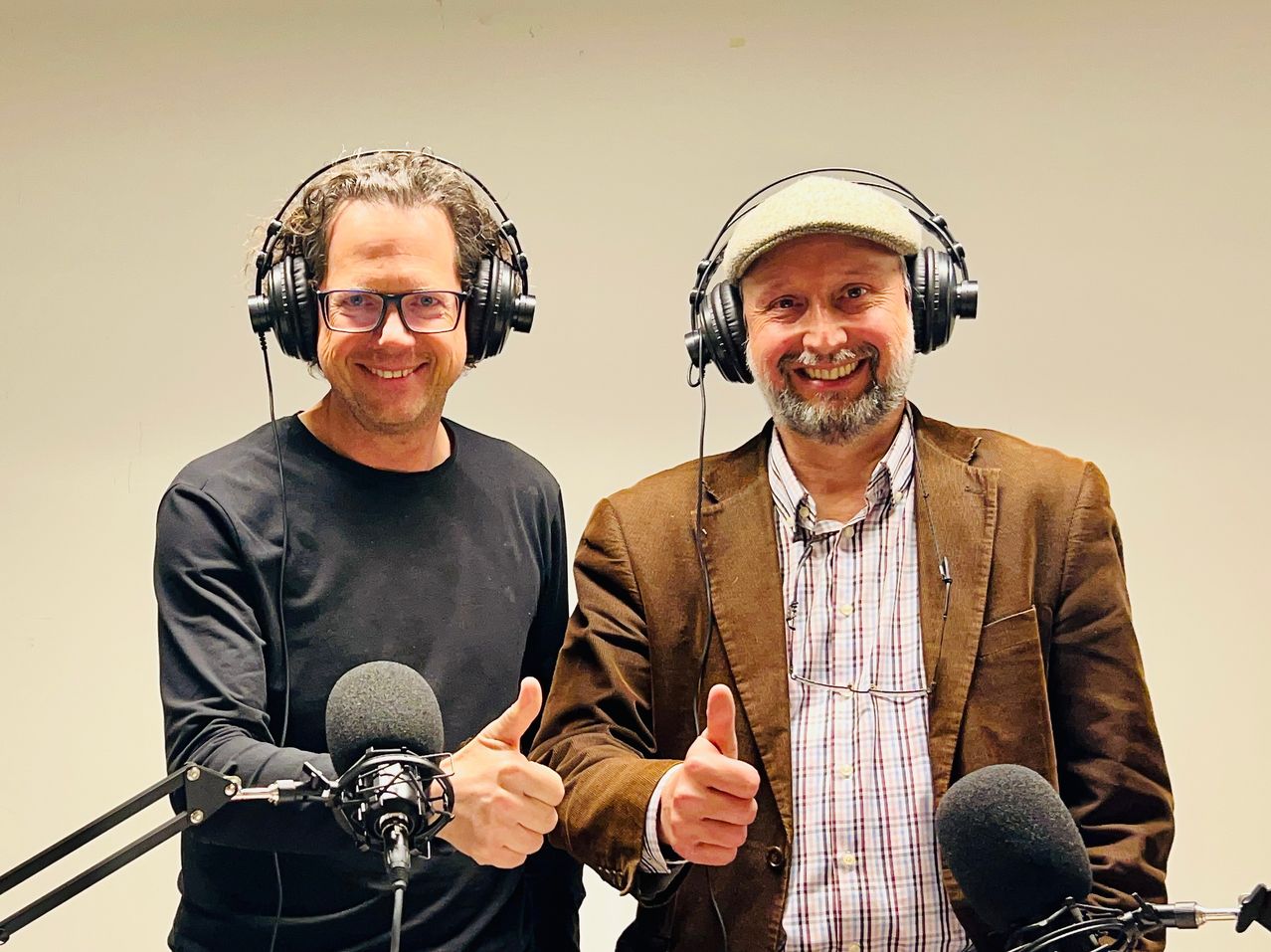 Zwei Männer in Kopfhörern geben vor Mikrofonen Daumen hoch. Ein Mann in einem schwarzen Shirt und der andere in einem braunen Jackett.