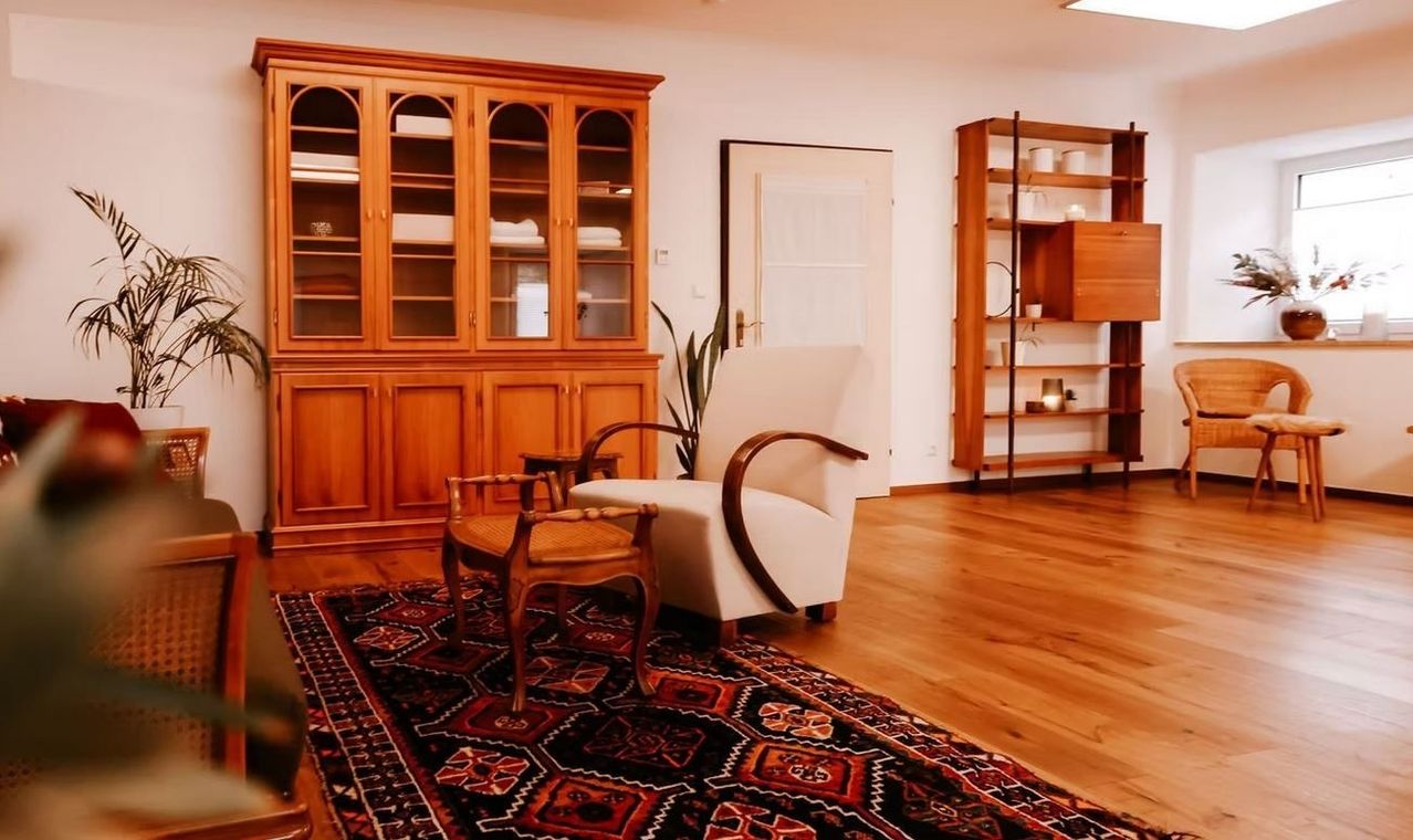 Ein Zimmer mit Holzböden hat einen großen hölzernen Schrank mit Glastüren, einen weißen Sessel und einen Holzstuhl. Ein Teppich liegt auf dem Boden. Eine Topfpflanze steht in der Ecke. Ein Regal mit Gegenständen ist auf der rechten Seite.