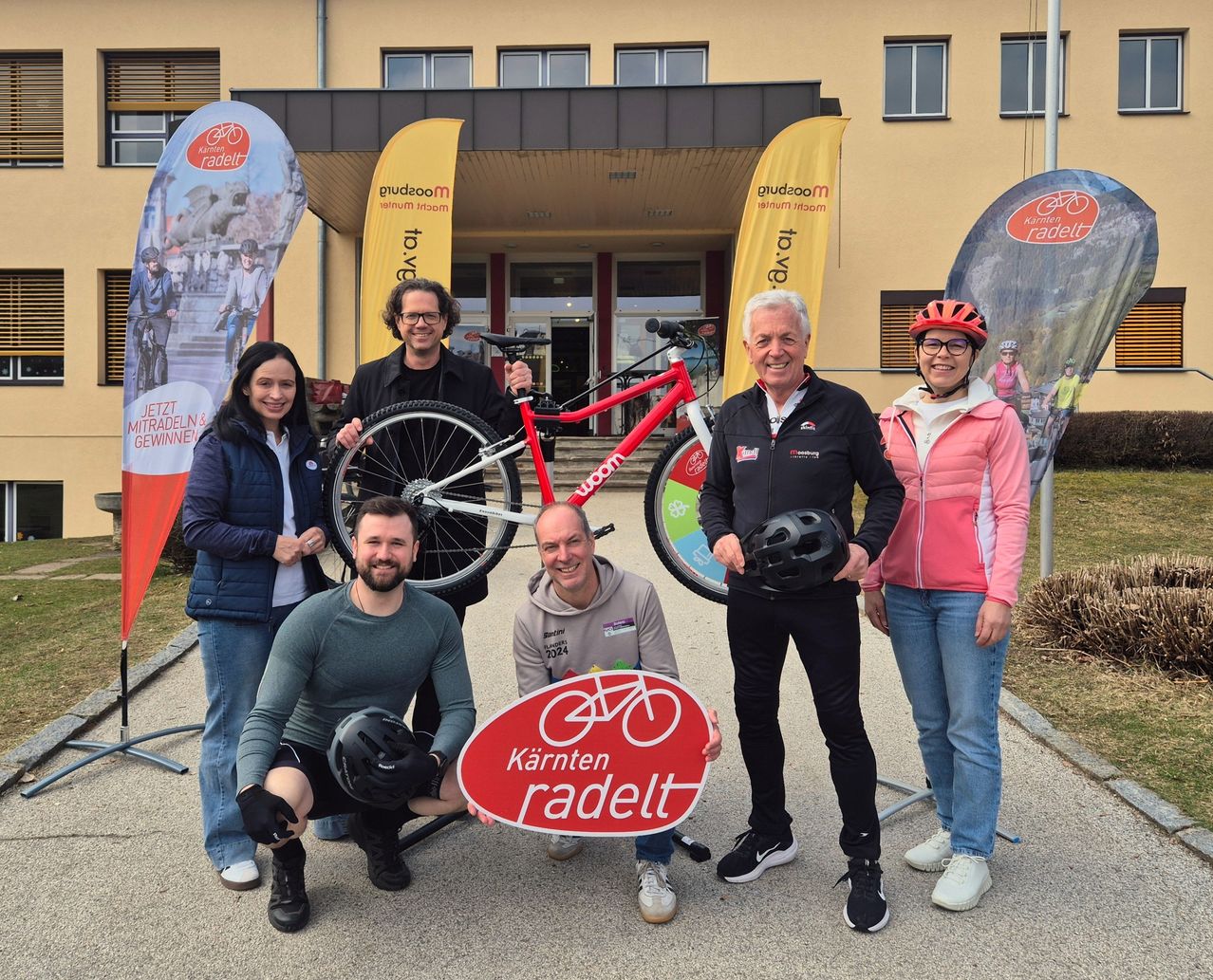 Eine Gruppe von Menschen posiert vor einem Gebäude mit Bannern, einer hält ein Fahrrad. Sie tragen alle Fahrradausrüstung, und der Mann rechts trägt einen roten Helm.