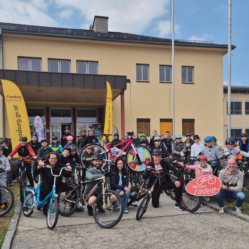 Eine Gruppe von Menschen posiert mit ihren Fahrrädern vor einem Gebäude. Einige tragen Helme. Ein Schild steht 'Kärnten radelt'. Das Gebäude hat einen Schornstein, Fahnen und Fenster.