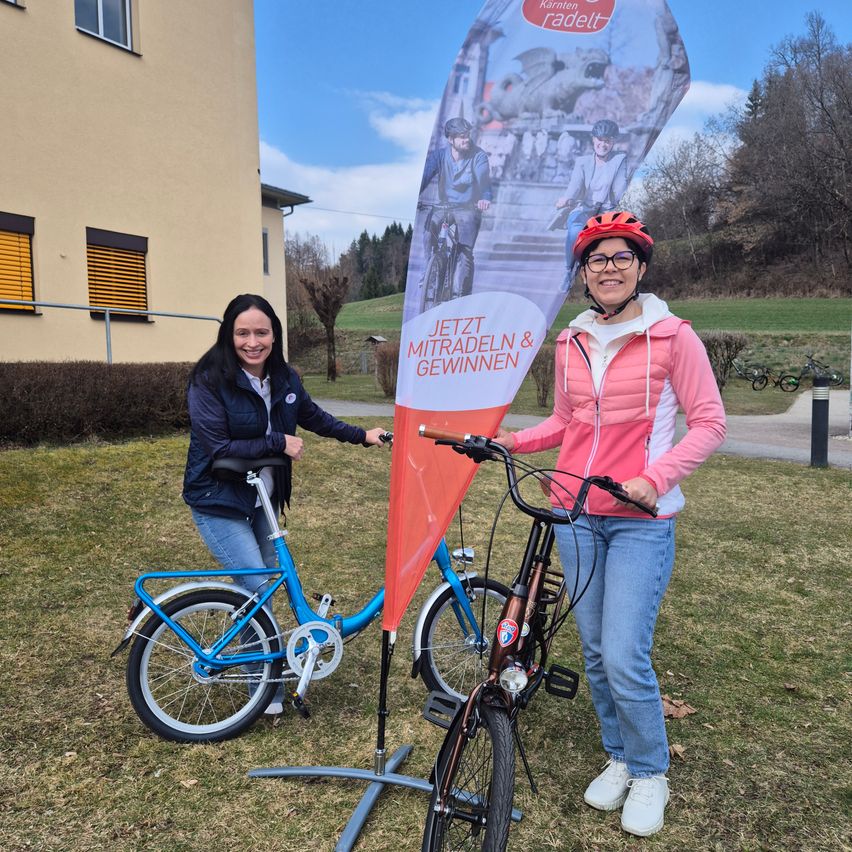 Zwei Frauen stehen mit ihren Fahrrädern vor einem Gebäude. Sie tragen Winterkleidung und Helme. Hinter ihnen ist ein Banner mit einem Bild von Radfahrern.